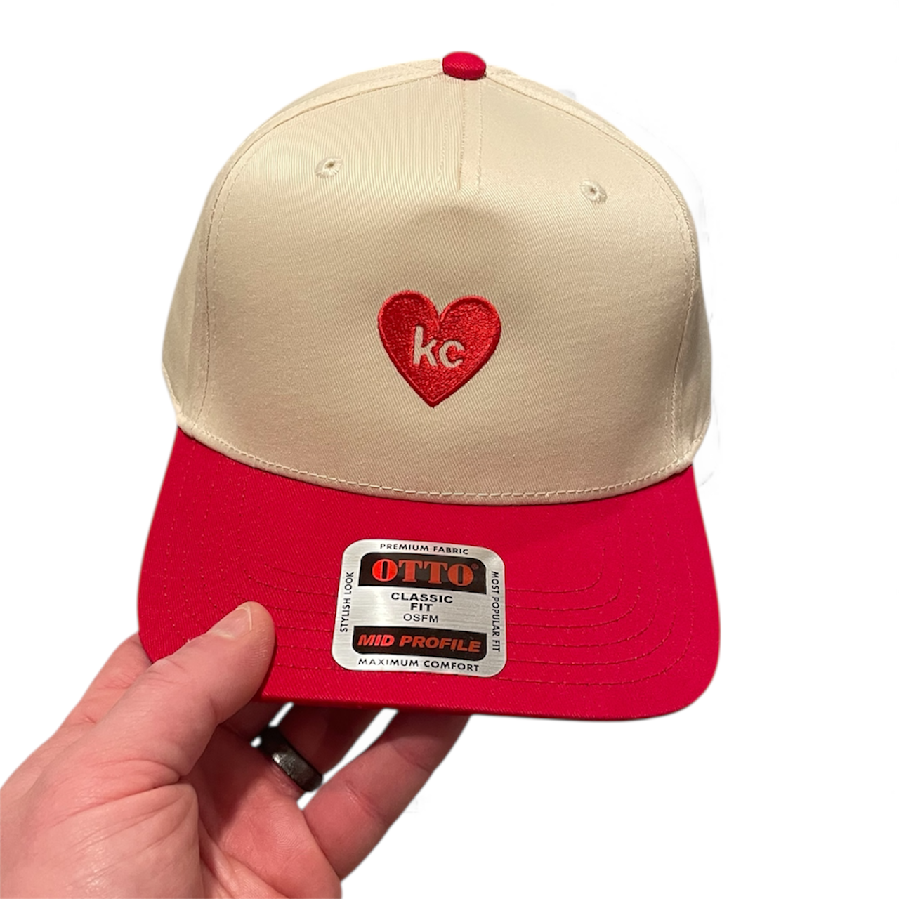 Beige and red cap with a heart-shaped logo held by a hand on a white background