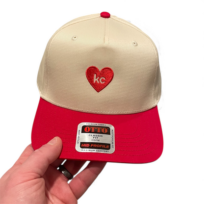 Beige and red cap with a heart-shaped logo held by a hand on a white background